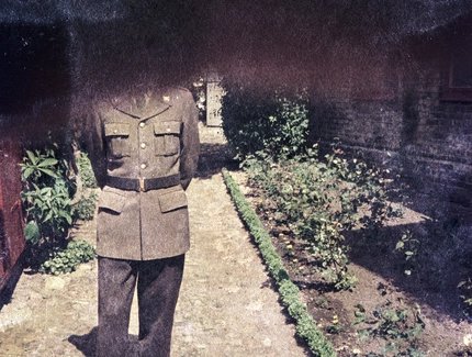 Fotografie: Farbnegativ, das eine Person in Uniform auf einem von Blumenbeeten gesäumten Weg zeigt. Der Kopf der Person und die ganze obere Hälfte der Fotografie sind dunkel angelaufen und dadurch verdeckt.
