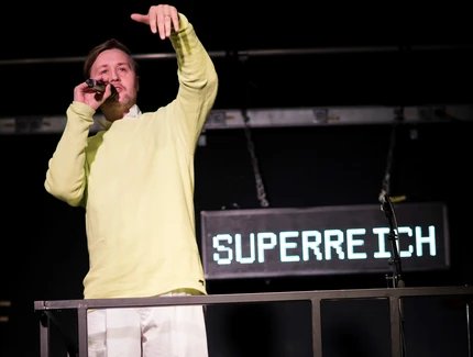 Eine Person in einem gelben Pulli und mit einer weissen Hose redet in ein Mikrophon. Hinter ihm hängt ein Schild auf dem Superreich steht.