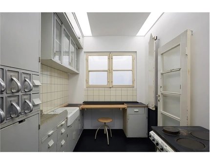 Raumfoto einer Küche mit Herd, Glastür, zugeklapptem Bügeltisch auf der rechten Seite; Schütten für unverpackte Waren aus Metall, Waschbecken und Lagerflächen auf der linken Seite. Im Hintergrund ist eine Arbeitsfläche mit Hocker, Schubladen und Fens