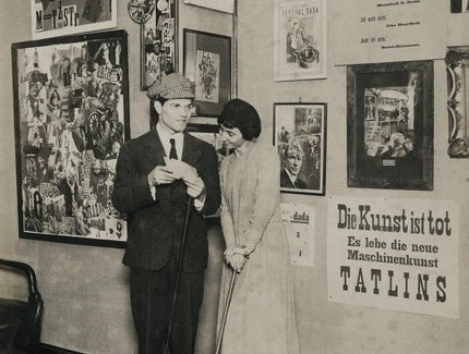 Schwarz-weiß-Foto von Raoul Hausmann und Hannah Höch. Die beiden stehen nebeneinander vor einer Wand voller gerahmter Werke und Poster.
