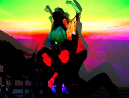 Eine Gruppe von fünf Performer:innen posiert in einer dynamischen, vielschichtigen Komposition. Sie sind vor einer Projektion, die einen leuchtenden Stadthimmel im Sonnenuntergang mit einem Farbverlauf zeigt. Die Projektion wirft starke, gesättigte F