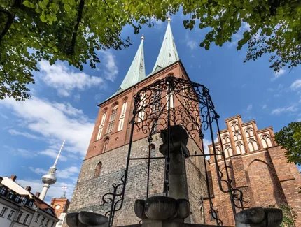 Blick durch eine schmiedeeiserne Brunnenzier auf die Türme und Giebel der Nikolaikirche, im Hinterfgrund hinter den Dächern historisierender Gebäude der Fernsehturm und der Turm des Roten Rathauses