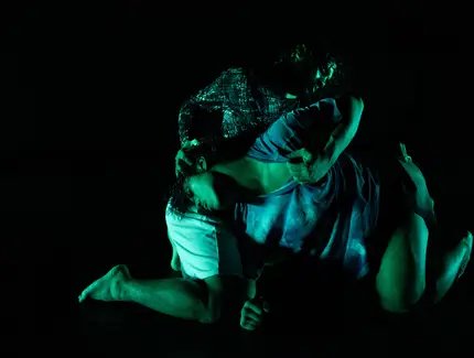 Zwei Performer:innen in intensiver Pose auf dunkler Bühne, grünlich beleuchtet, mit ineinander verschlungenen Körpern.