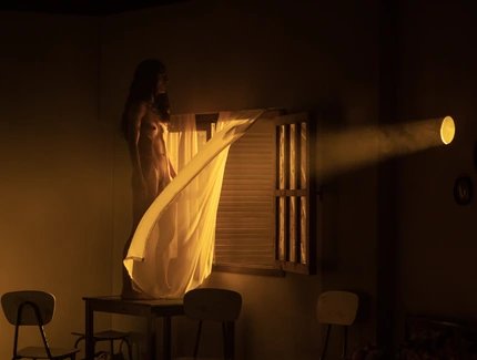 Eine nackte Frau steht auf einem Tisch vor einem offenen Fenster durch das von rechts das Licht eines Scheinwerfers scheint.