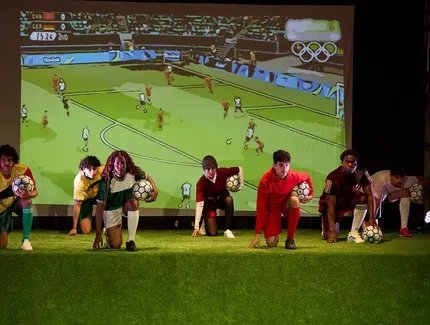 Eine Gruppe Menschen kniet vor einer Projektion mit jeweils einem Fußball unter dem Arm.