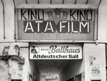 Junge auf Tretroller vor Toreinfahrt in einen Hinterhof, darüber deutsch- und türkischsprachige Werbung