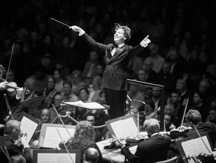 Dirigent Klaus Mäkelä breitet die Arme vor dem Orchester aus, die Musiker:innen streichen ihre Geigen und im Hintergrund sieht man das Publikum.