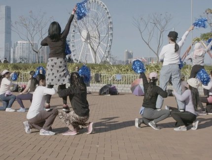 Video-Still: Eine Gruppe von Menschen im Freien mit blauen Pompons, einige kniend und andere stehend, vor einem Riesenrad und Hochhäusern in einer Stadtlandschaft.