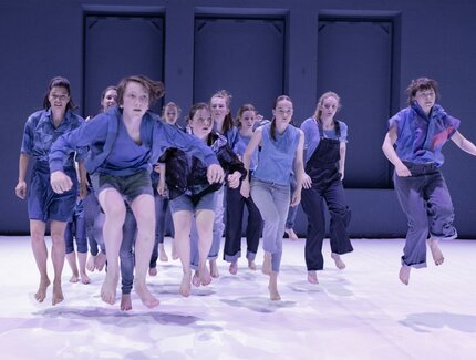 Eine Gruppe blaugekleideter junger Menschen vollzieht eine Sprungbewegung auf einer Bühne.