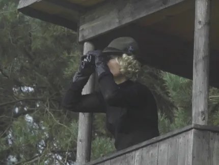 Ein blonde Frau beobachtet auf einem Hochsitz im Wald etwas mit einem Fernglas.