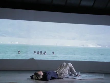 Zu sehen ist eine Person, die vor einer Videoprojektionsleinwand auf dem Boden liegt. Es wird eine Aufnahme im Ultabreitformat von in einem Gewässer badenden Menschen projiziert. Die Person, die auf dem Boden liegt, hat ihre Beine aufgestellt und ihr