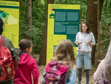 Eine Frau erzählt vor einem Schild stehend die Geschichte der mittelalterlichen Siedlung im Museumsdorf Düppel