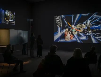 Foto: Ein paar Menschen sitzen und stehen in einem Videoraum und schauen auf eine große Leinwand.