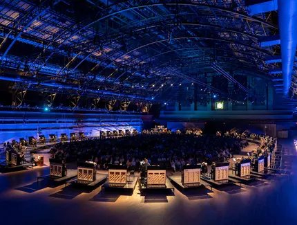 In einer großen Halle formen zahlreiche Klaviere eine Ellipse um das Publikum herum.