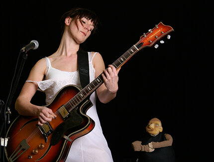 Rike Schuberty in weißem Unterkleid mit großer E-Gitarre vor einem Mikrofon. Im Hintergrund sind eine kleine Puppe aus Schaumstoff.