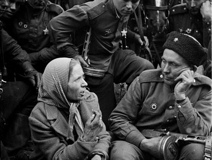 Mehrere Soldaten in Uniform sitzen und stehen neben einem Pferd, eine ältere Frau mit Kopftuch sitzt darunter