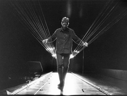 Es sind eine Reihe von Saiten quer durch einen Raum gespannt. Dazwischen steht eine Person und zupft sie.