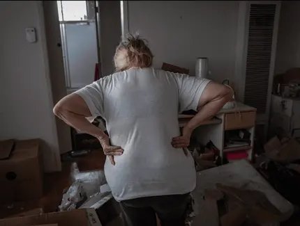 Das Foto zeigt eine Frau, die mit dem Rücken zur Kamera in einem unordentlichen, chaotischen Raum steht. Sie trägt ein weißes T-Shirt und dunkle Hosen und stützt ihre Hände in den unteren Rücken, als sei sie erschöpft oder habe Schmerzen.