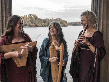 Die Musikerinnen des Ensembles Triphonia mit Instrumenten in einem Pavillion am See stehend, miteinander scherzend