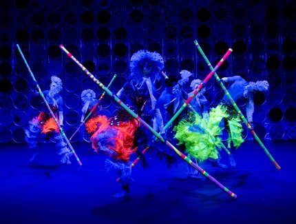 Die rüschigen Kostüme und langen Stäbe von den Tänzer:innen leuchten in bunten Neonfarben.