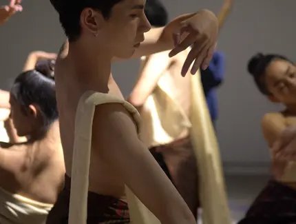 Mehrere Performer:innen mit nacktem Oberkörper und traditionell anmutenden Stoffbahnen in sanften, fließenden Bewegungen; Fokus auf eine zentrale Person mit erhobenen Armen in meditativer Pose.