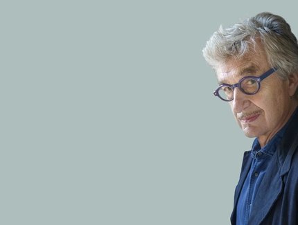 Ein Mann mit grauen Haaren und blauer Brille schaut schmunzelnd aus dem rechten Bildrand in die Kamera.