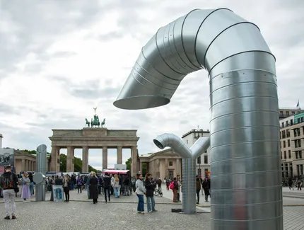 Zwei große, gebogene Metallstrukturen stehen auf dem Pariser Platz vor dem Brandenburger Tor. Menschen versammeln sich in der Nähe.