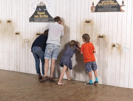 Eine vierköpfige Familie steht vor einer weißen Wand. Durch Gucklöcher in der Wand werfen sie einen Blick auf dahinter angebrachte Fotografien historischer Flugplätze. Auf der Wand sind historische Werbeplakate für Wettflüge aufgehängt.
