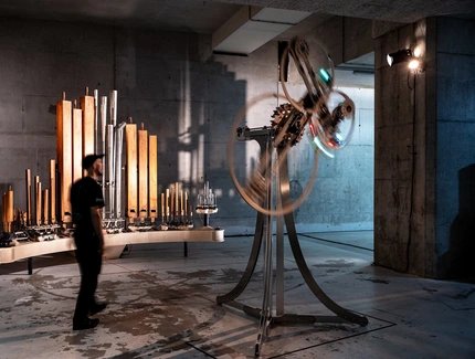 In einem kargen Raum stehen zwei Objekte. Eines besteht aus unterschiedlichen, nebeneinander aufgereihten Orgelpfeifen, das andere aus einem Stativ und sich drehenden Kreisen.