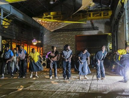 Eine Gruppe von Menschen tanzt energisch in einem großen Veranstaltungsort im Industriestil, der von bunten Lichtern und Discokugeln beleuchtet wird, mit Zuschauern im Hintergrund.