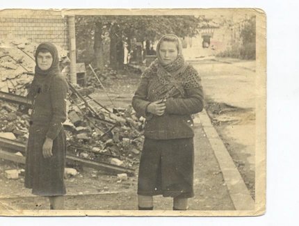 Das Foto zeigt zwei junge Mädchen vor dem Hintergrund von Trümmern. Das Mädchen links trägt ein dunkles Kleid und ein Kopftuch. Das Mädchen rechts trägt ein dunkleres Kleid, Strümpfe und einen gehäkelten Schal. Der Hintergrund deutet auf Zerstörung u