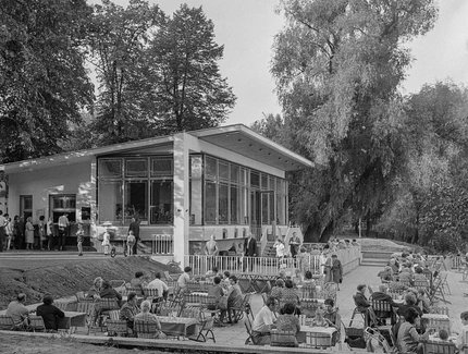Schwarz-Weiß-Fotografie des Milchhäuschens in Berlin-Weißensee, vor dem viele Menschen in kleinen Grüppchen an Tischen im Außenbereich sitzen. Das Milchhäuschen wird von hohen Bäumen gesäumt.