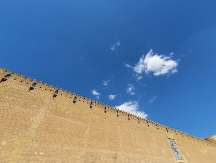 Das Bild zeigt eine hohe mit schmuckvollen Zinnen versetzte Mauer, die gegen einen strahlend blauen Himmel mit wenigen Schönwetterwolken fotografiert ist.