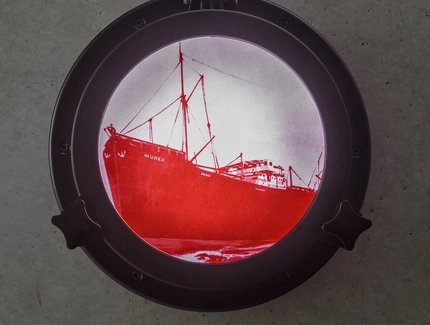 Foto: Blick auf ein rundes Fenster (Bullauge), in dem in leuchtend roter Farbe auf weiß-rosa Hintergrund ein großes Schiff zu sehen ist. Am Bug des Schiffes steht in Großbuchstaben 'Murex'.
