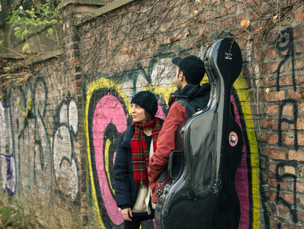 Zwei Personen stehen vor einer Backsteinwand, die mit Graffiti besprayt ist. Bei der linken Person handelt es sich um die Gastgeberin Antonia Regis. Die rechte Person ist nur von hinten zu sehen, sie trägt ein Cello-Koffer.