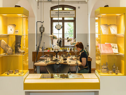 Zwischen zwei gelben Vitrinen mit unterschiedlichen Objekten blickt man auf einen Tisch mit zahlreichen, verschiedenen Goldschmiede-Geräten. An dem Tisch sitzt eine Frau und arbeitet.