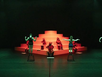 Performer:innen auf Hockern und Musiker:innen in roten Kapuzen posieren vor einer stufenförmigen weißen Plattform im grünen und roten Licht.