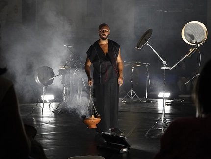 Ein Mann in einem ärmellosen schwarzen Outfit steht auf einer dunklen, rauchigen Bühne und hält einen Ton-Räuchergefäß in der Hand, aus dem Rauch aufsteigt. Um ihn herum stehen unter sanftem weißem Licht Percussion-Instrumente und Mikrofone, während 