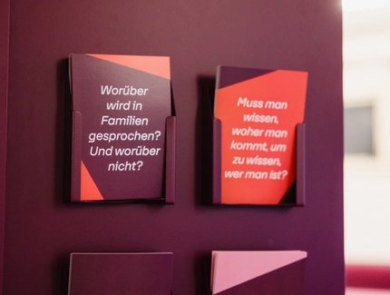 Zwei Informationskarten in Haltern an einer dunkelroten Wand, eine dunkelrot mit weißem Text 'Worüber wird in Familien gesprochen? Und worüber nicht?', die andere rot mit weißem Text 'Muss man wissen, woher man kommt, um zu wissen, wer man ist?'.