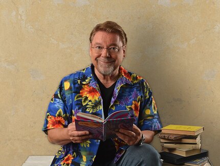 Der Autor und Vorleser mit einem offenen Buch in den Händen, auf einem Palettenstapel sitzend mit einem Bücherstapel neben sich.