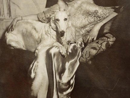 Selbstportrait der Künstlerin Marta-Astfalck-Vietz als Sepia-Fotografie. Sie sitzt in einem glänzenden Kleid mit Pelzbesatz auf einem Sofa. Auf ihrem Schoß sitzt ein Hund mit hellem Fell. Im Hintergrund sind Gemälde und dekorative Elemente zu sehen.