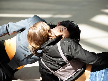 Zwei Personen liegen einander versetzt gegenüber auf dem Boden in der Sonne. Ihre Münder berühren sich.