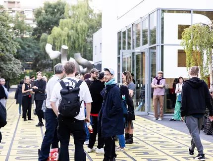 Auf dem Vorplatz der Berlinischen Galerie stehen einige Menschen in kleineren Gruppen zusammen und unterhalten sich miteinander. Eine Person blickt direkt in die Kamera.