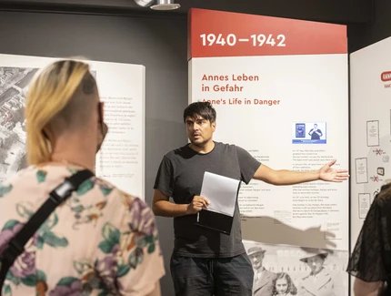 Ein Mann erklärt vor einer Ausstellungswand mit historischen Informationen einer kleinen Gruppe etwas und gestikuliert dabei.
