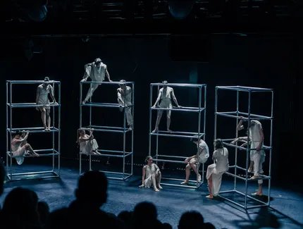 Auf einer dunklen Bühne stehen vier silberne Gerüste nebeneinander, auf denen sich mehrere Tänzer*innen stehend oder sitztend positionieren. Sie tragen weiße, schlichte Kostüme.