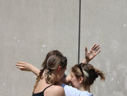 Zwei junge Frauen stehen dicht beieinander und verschränken ihre Arme ineinander.