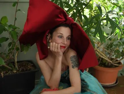 Meret Becker sitzt in einem Kleid aus türkis- und rosa-farbenem Stoff mit einer großen roten Schleife auf dem Kopf in der Hocke. Der Hintergrund zeigt verschiedene Zimmerpflanzen.