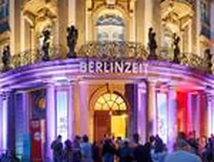 Das Museum bei Nacht von außen mit bunter Leuchtschrift, auf der BERLINZEIT steht, davor Menschen