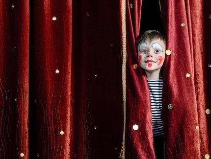 Kind als Clown geschminkt, hinter einem roten Vorhang 