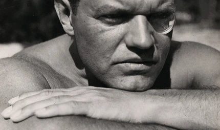 Schwarz-Weiß-Fotografie von Raoul Hausmann, der mit runder Sehhilfe im linken Auge, den Kopf auf den verschränkten Armen aufgelegt, an den Betrachtenden vorbei in die Ferne blickt.